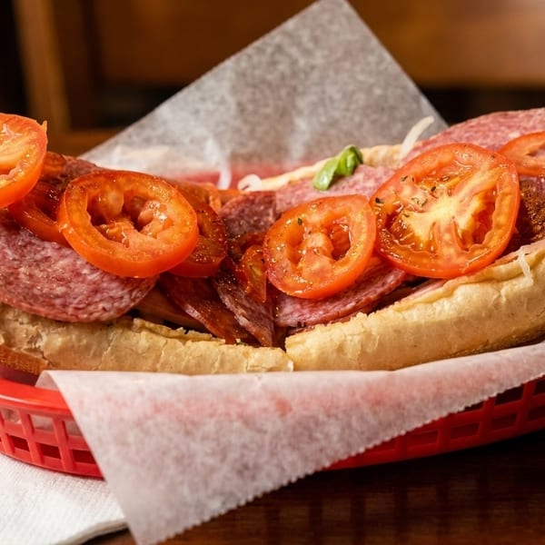 A large deli sub sandwich with salami, ham, and sliced tomatoes in a red basket.