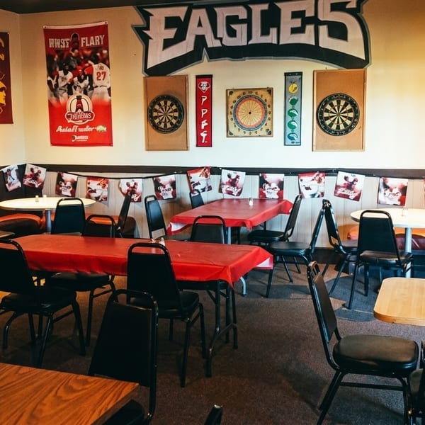 Casual restaurant interior with tables, chairs, sports banners, and dartboards on the walls.