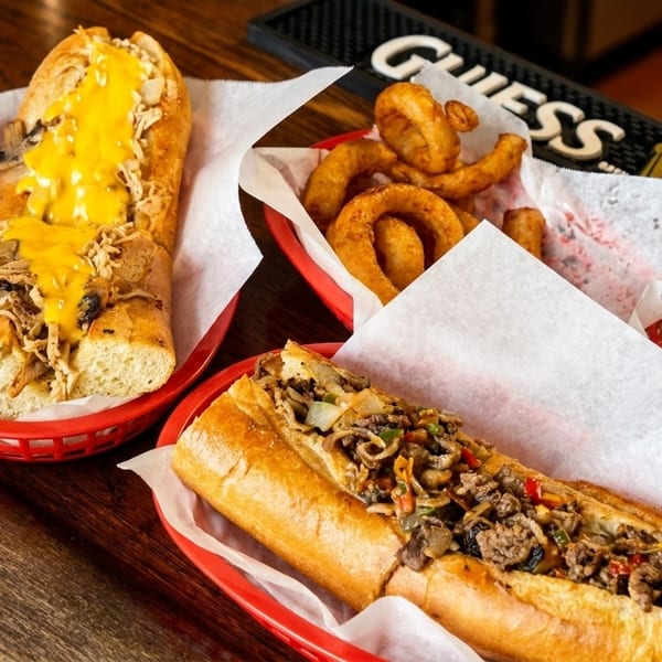 Two cheesesteak sandwiches and a basket of onion rings on a wooden table with a Guinness bar mat.